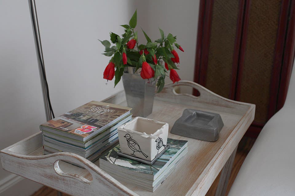 En primer plano, junto al sillón, bandeja con patas a modo de mesa lateral. Sobre ella, florero, cenicero, macetita ''chorreada ilustrada'' de loza ($65, Artefactos) y una colección personal de libros y revistas de deco y diseño