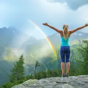 Una mujer en la montaña levantando los brazos con un arcoíris de fondo