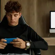 Adolescente con celular en la mano y computadora de fondo.