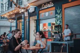 Dos chicas sentadas en una mesa en la vereda tomando y comiendo.