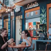 Dos chicas sentadas en una mesa en la vereda tomando y comiendo.