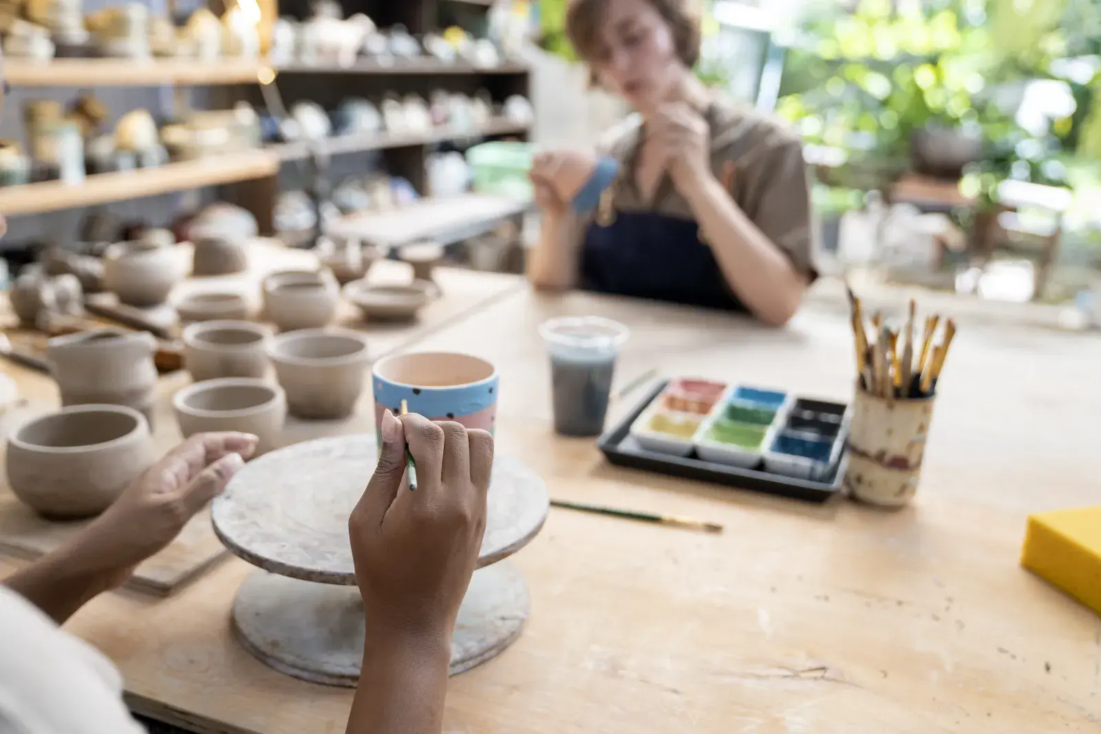 Manos de mujer decorando una pieza de cerámica y una mujer en segundo plano pintando una cerámica.