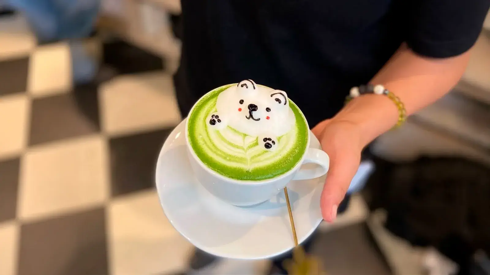 Plano medio de una persona sosteniendo un plato con una taza de café. En ella se encuentra un café de color verde con el relieve de espuma con forma de oso tipo kawai