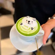Plano medio de una persona sosteniendo un plato con una taza de café. En ella se encuentra un café de color verde con el relieve de espuma con forma de oso tipo kawai
