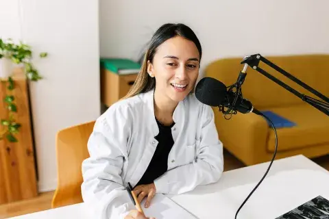 Una mujer hablando por micrófono, haciendo un podcast desde su casa, mientras toma nota Una mujer hablando por micrófono, haciendo un podcast desde su casa, mientras toma nota