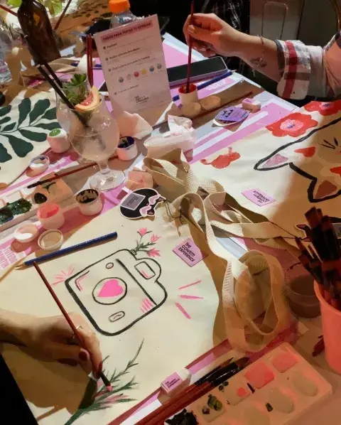 Elementos sobre una mesa para pintar una totebag y chicas pintando.