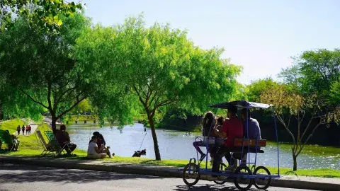 Termas de General Belgrano, un lugar donde se ve un espejo de agua y un espacio verde con gente descansando. En primer plano, un carrito de paseo con cuatro personas pedaleando. Termas de General Belgrano, un lugar donde se ve un espejo de agua y un espacio verde con gente descansando. En primer plano, un carrito de paseo con cuatro personas pedaleando.