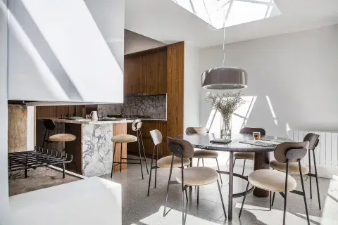 La cocina con isla + mesa redonda cercana. Se ven las banquetas en la isla y las sillas haciendo juego con la mesa. Lámpara ‘Pluvial’ de Federico Churba corona una mesa ‘Estribo’, de La Feliz. Alrededor, las sillas ‘42′ de la misma marca. Paredes y muebles (Estudio Ordóñez-Wenzke) enchapados en palisandro. Mesadas y alzadas en cuarcita.