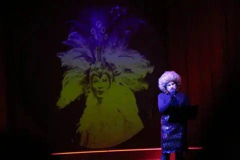 Juan Pablo Geretto vestido de mujer con una peluca y detrás una imagen de otra mujer con plumas. Juan Pablo Geretto vestido de mujer con una peluca y detrás una imagen de otra mujer con plumas.