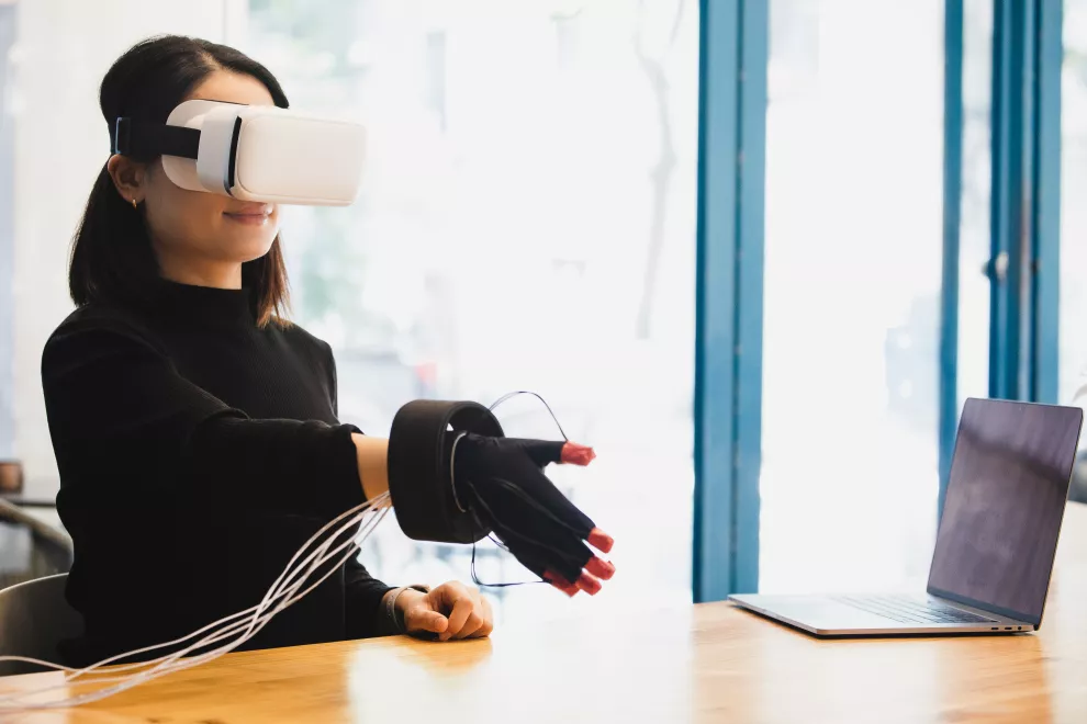 Una mujer con casco de realidad virtual y guante háptico interactuando con una persona/objeto virtual en el metaverso.