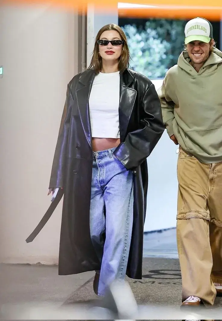 Hailey Bieber con crop top que deja ver su panza y loose jeans. El trench de cuero cierra un look casual y cool. 