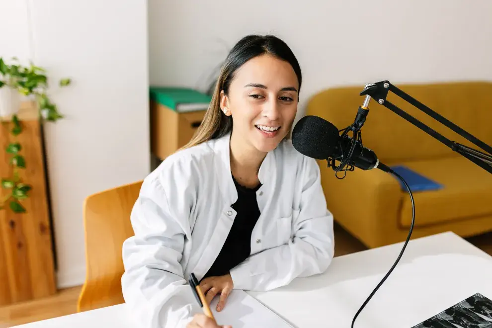 Una mujer hablando por micrófono, haciendo un podcast desde su casa,  mientras toma nota