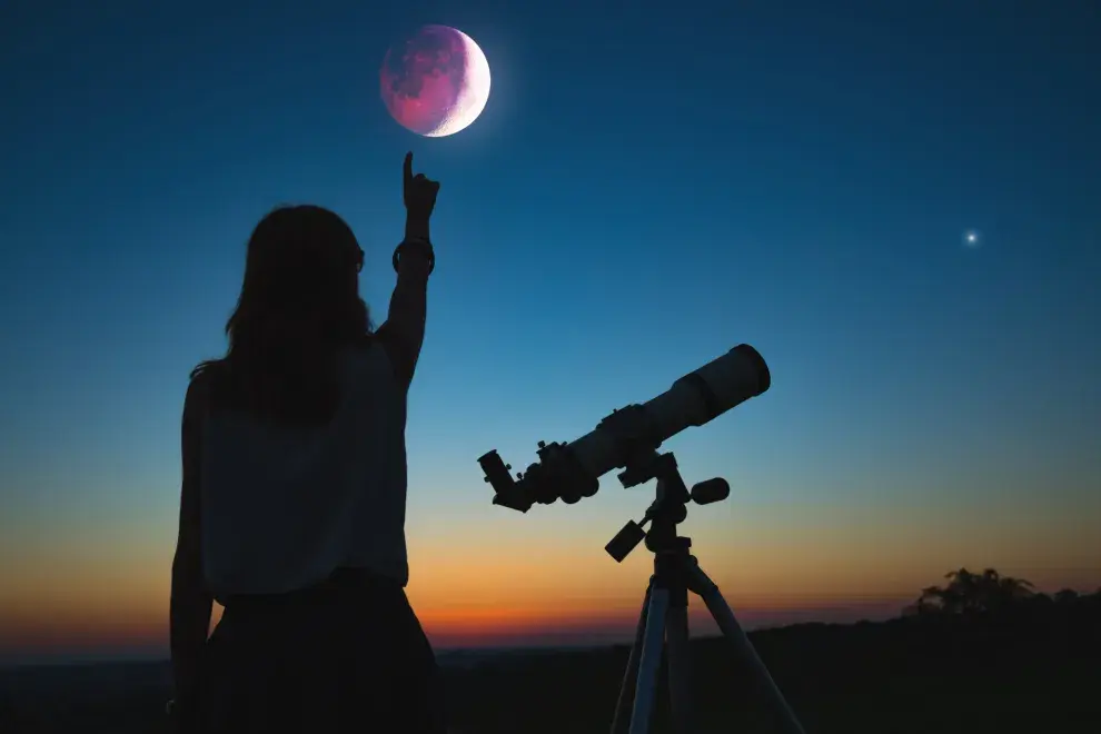 Mujer mirando una luna llena con un telescopio al costado.