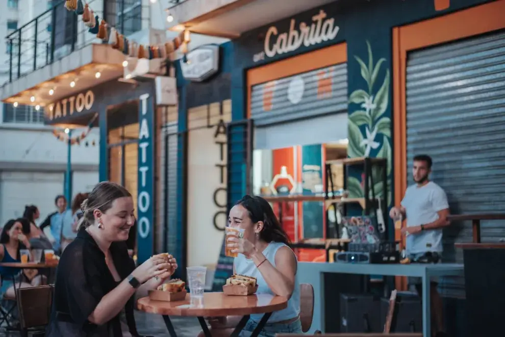 Dos chicas sentadas en una mesa en la vereda tomando y comiendo.