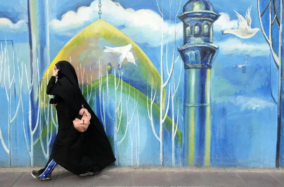 Una mujer cubierta de pies a cabeza camina frente a un mural en una mezquita del centro de Teherán, Irán,