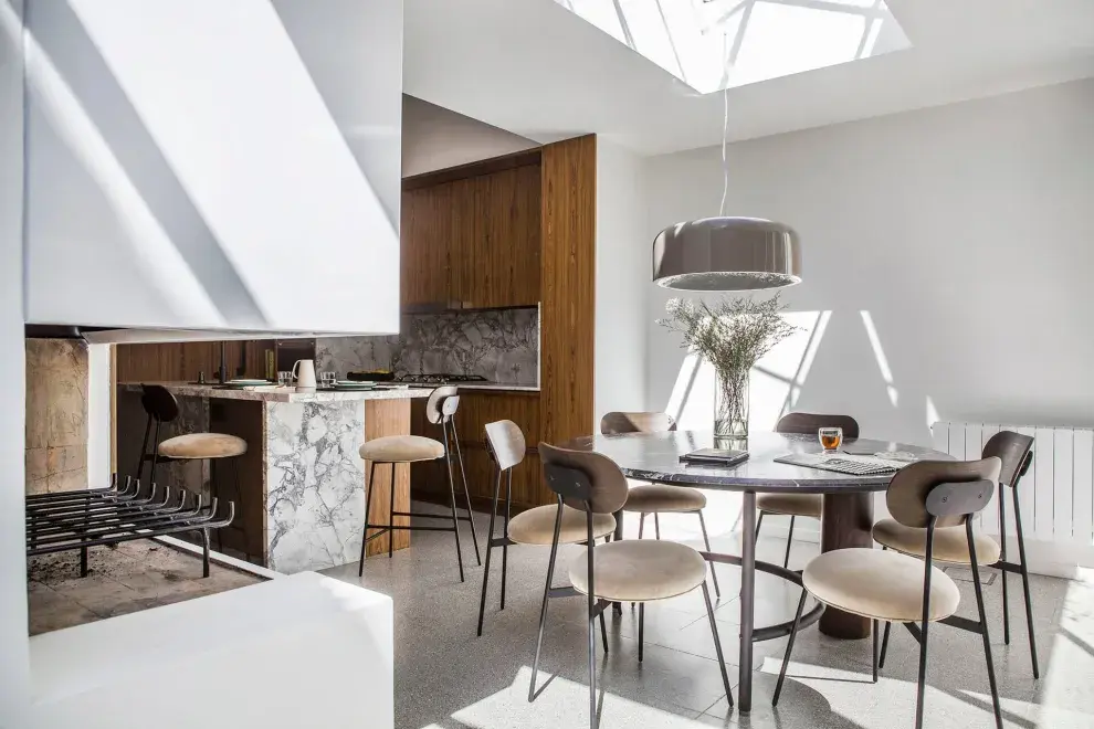 La cocina con isla + mesa redonda cercana. Se ven las banquetas en la isla y las sillas haciendo juego con la mesa. Lámpara ‘Pluvial’ de Federico Churba corona una mesa ‘Estribo’, de La Feliz. Alrededor, las sillas ‘42′ de la misma marca. Paredes y muebles (Estudio Ordóñez-Wenzke) enchapados en palisandro. Mesadas y alzadas en cuarcita.