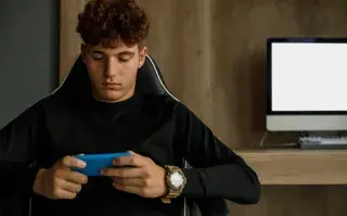 Adolescente con celular en la mano y computadora de fondo.