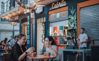 Dos chicas sentadas en una mesa en la vereda tomando y comiendo.
