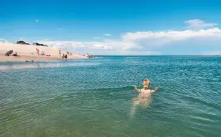 Fotografía de una chica nadando en un mar cristalino y de lejos se ve la arena blanca con un par de personas