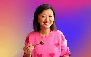 Recorte de Karina Gao sonriendo mientras sostiene unos palillos con un fondo multicolor amarillo, violeta y rosa perteneciente a la placa de OHLALAND!