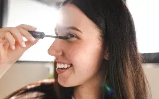 Una mujer pintándose las pestañas con una máscara frente al espejo