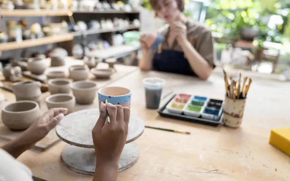 Manos de mujer decorando una pieza de cerámica y una mujer en segundo plano pintando una cerámica.