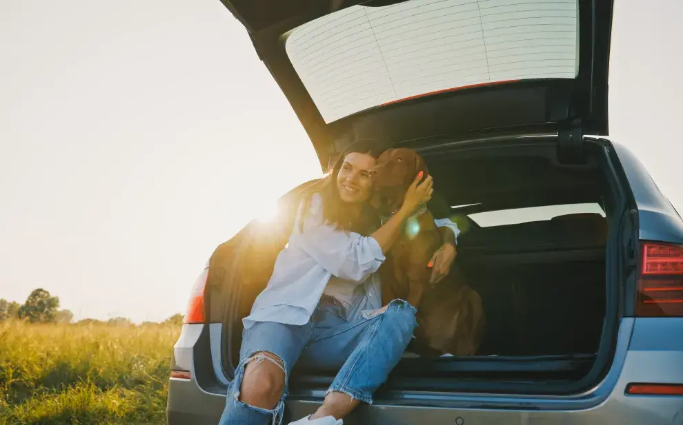 Una chica con su mascota sentadas en el baúl de un auto.